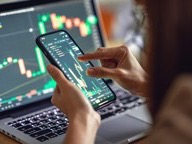 Person checking crypto price charts on a smartphone in front of a laptop